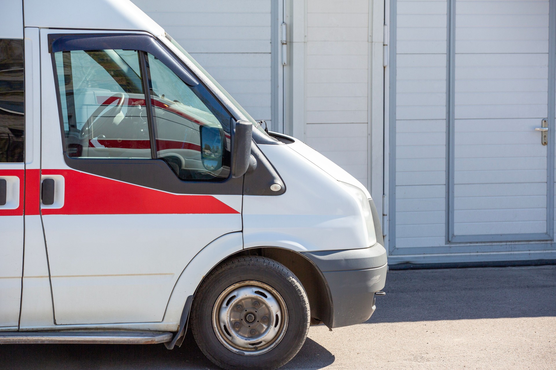 An ambulance is seen parked near the entrance of a modern building. The vehicle is white with a red stripe and equipped with blue emergency lights on top.